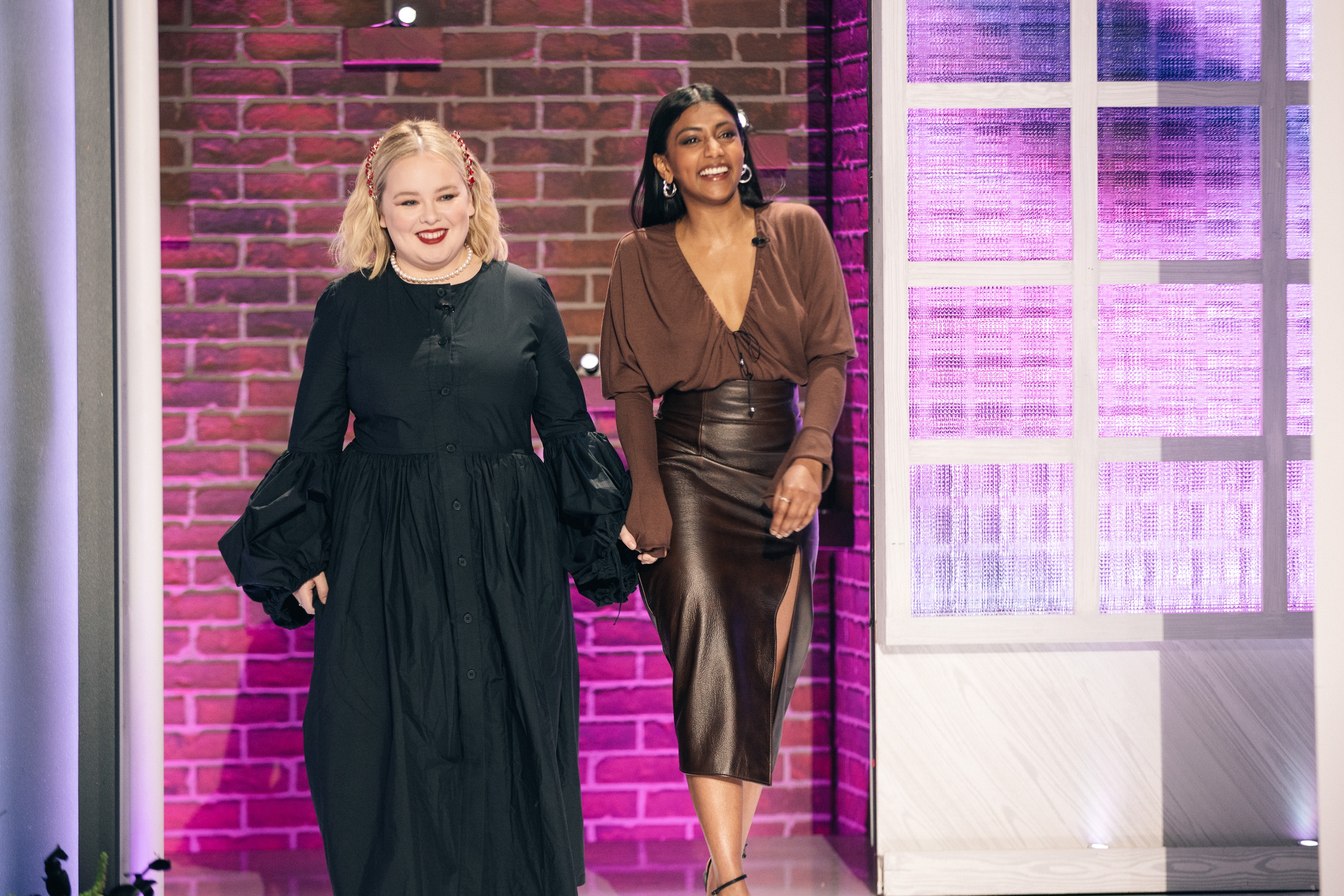 Two women, one in a long black dress and the other in a brown top with a leather skirt, walk hand-in-hand on a stage with a brick backdrop