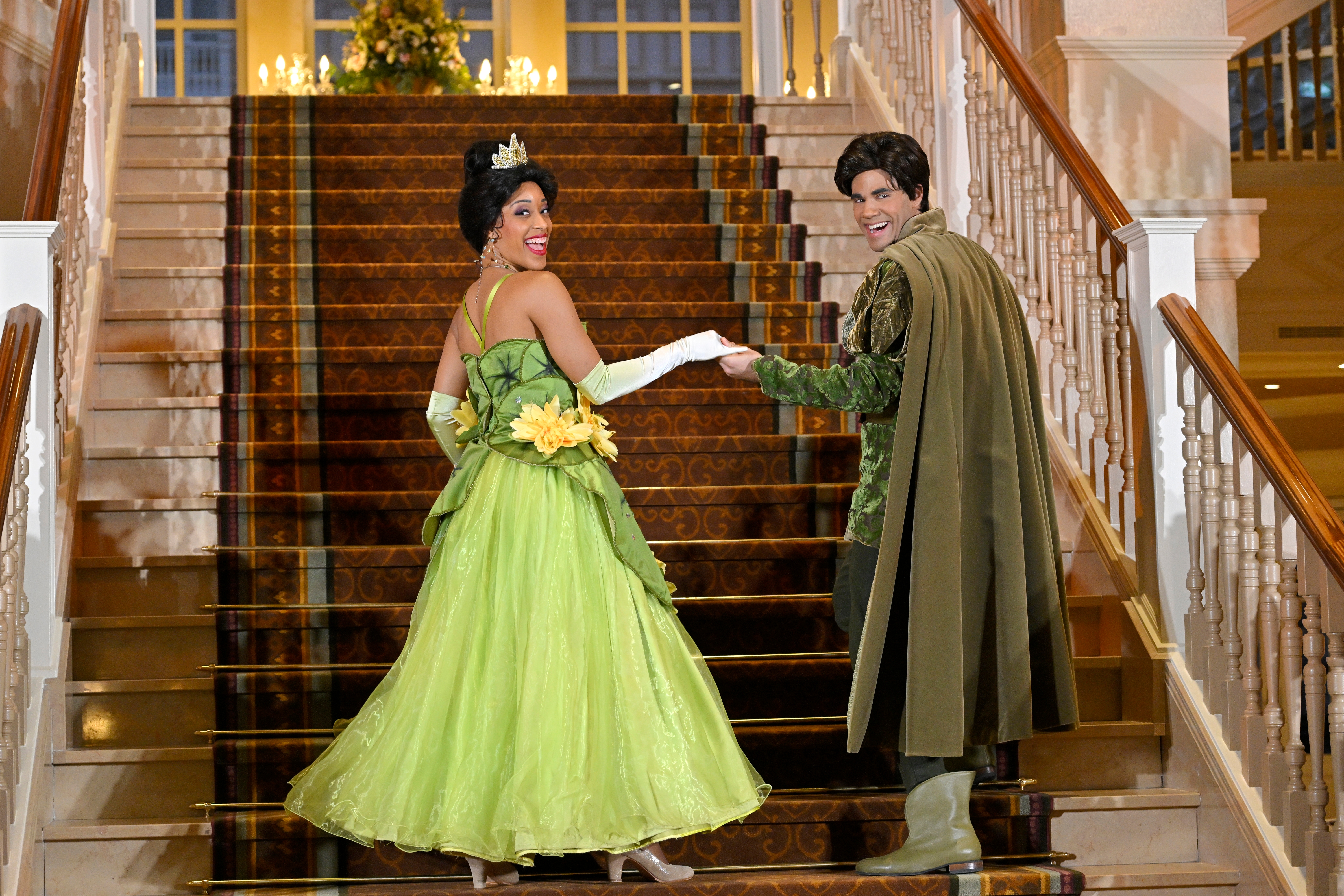 Two people in princess and prince costumes smile while standing on a staircase, holding hands, and looking back at the camera