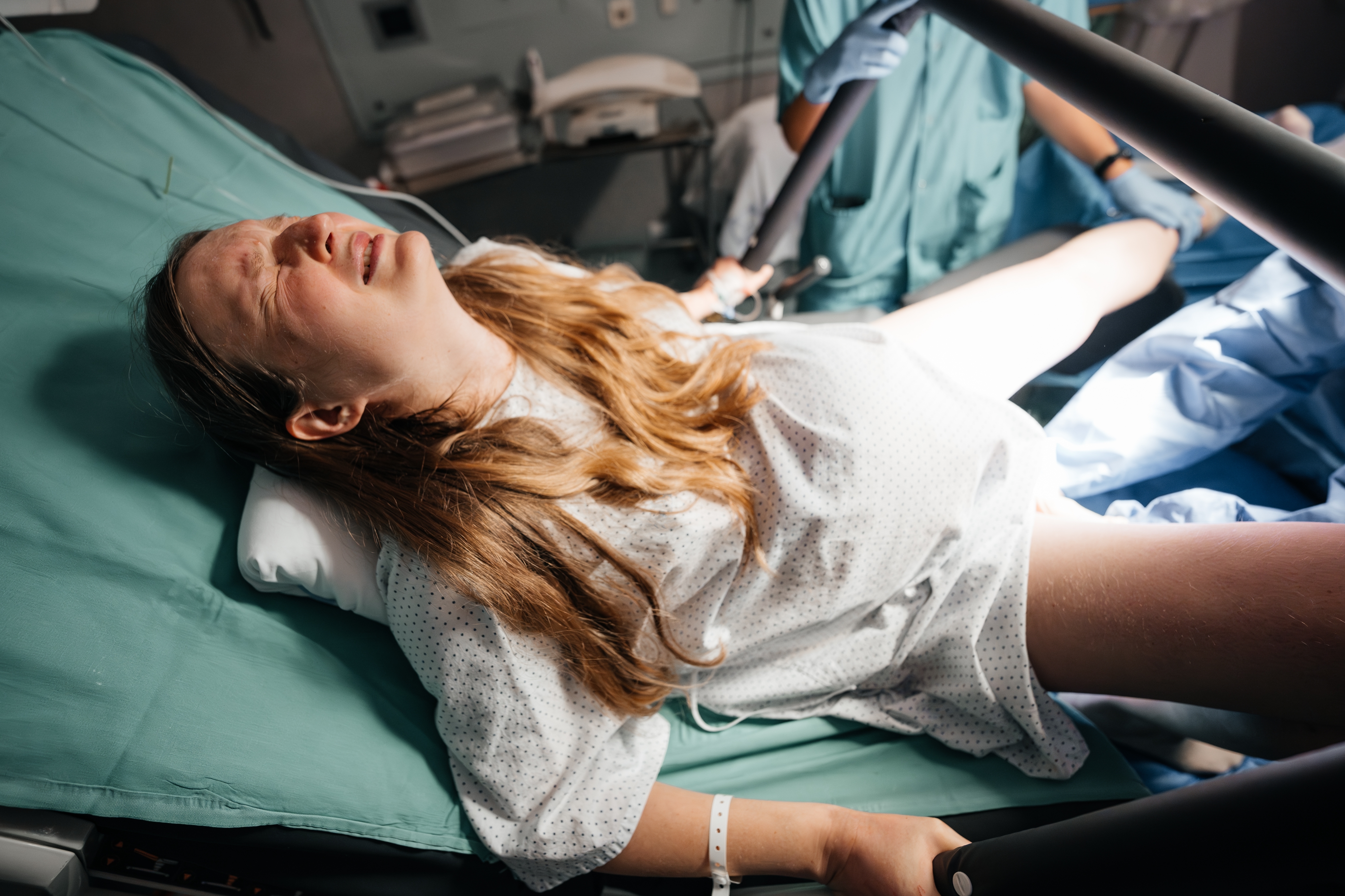A woman in a hospital bed appears to be in labor, gripping handles with a tense expression while a medical professional assists her