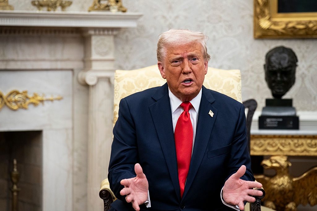 A person in a suit and red tie speaks with open hands in a formal room with historical decor