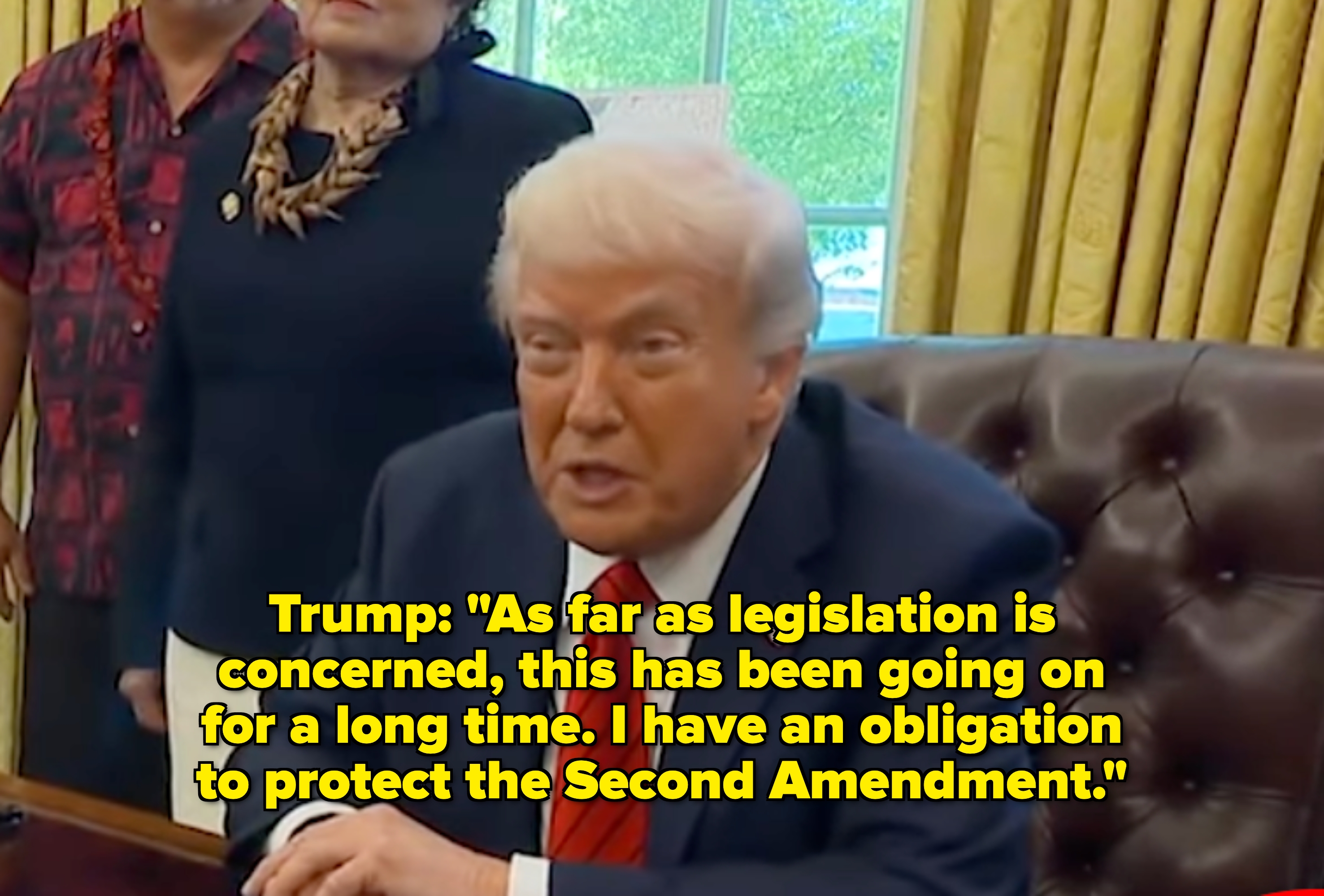 A person in a suit and red tie is sitting at a desk in an office setting, speaking, with two people standing in the background
