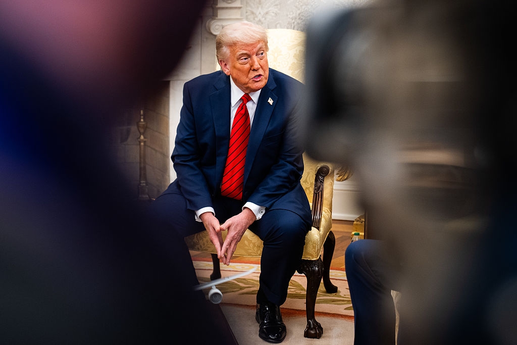 Person in a suit and red tie seated during an interview, framed by out-of-focus elements, suggesting a candid moment in a formal setting