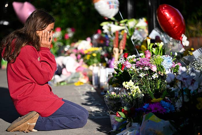Person kneels, covering face, at a memorial with flowers and balloons, reflecting somber mood