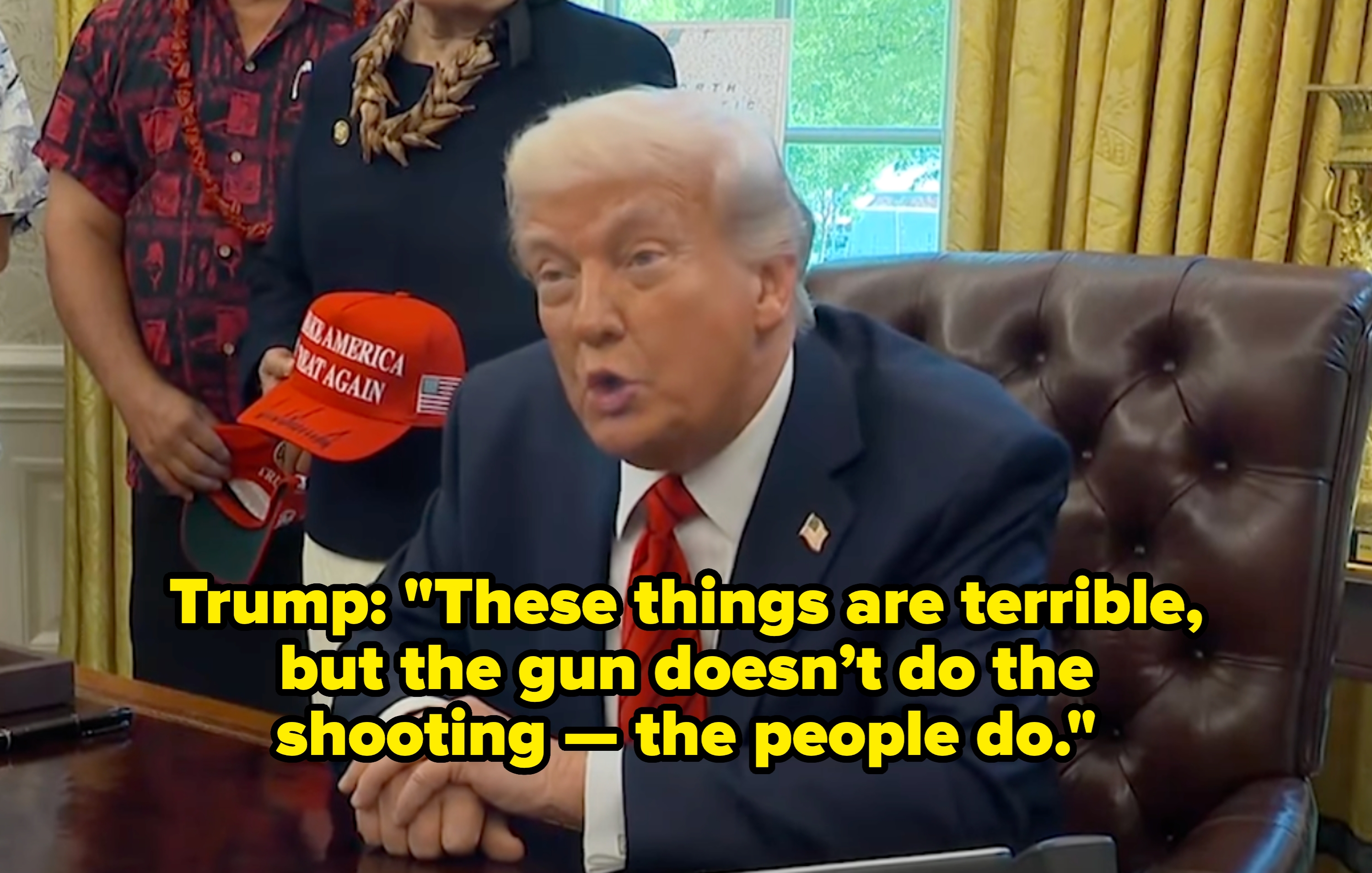 A man in a suit sits at a large desk in an office, talking. A person nearby holds a red hat with a slogan