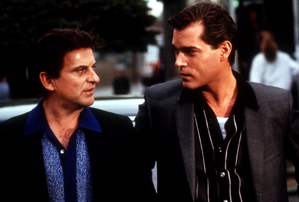 Two men in a scene from a movie, dressed in retro suits and shirts, engaged in an intense conversation outdoors