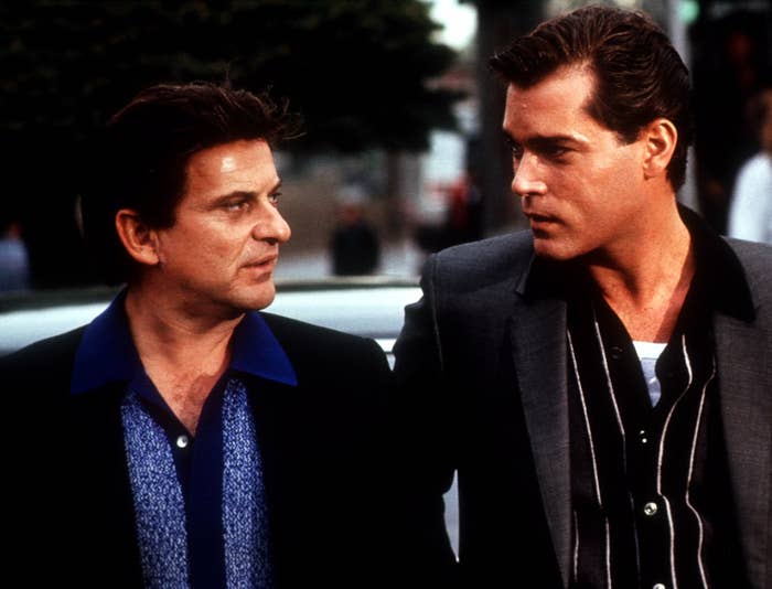 Two men in a scene from a movie, dressed in retro suits and shirts, engaged in an intense conversation outdoors