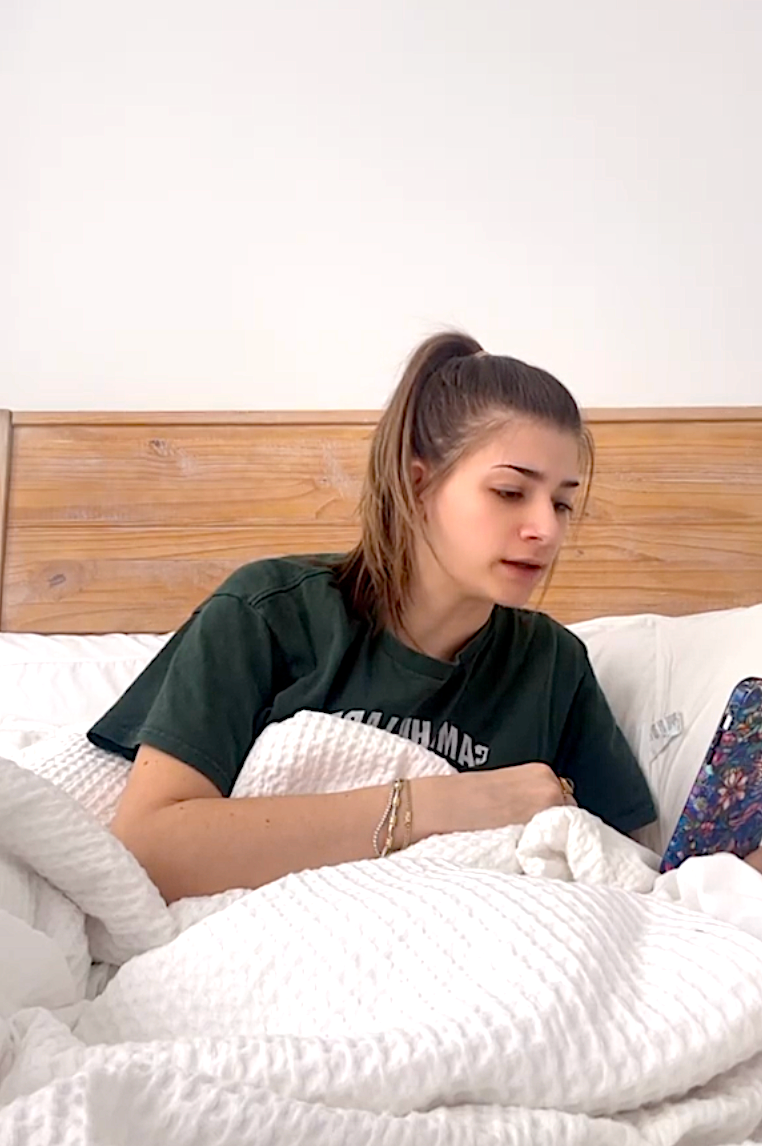 Person sitting in bed with a ponytail, looking at a phone while partially covered by a white blanket