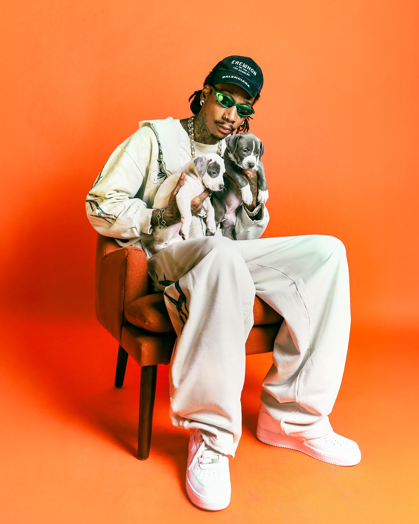 Person in relaxed outfit and cap sits on a chair holding two puppies against an orange backdrop
