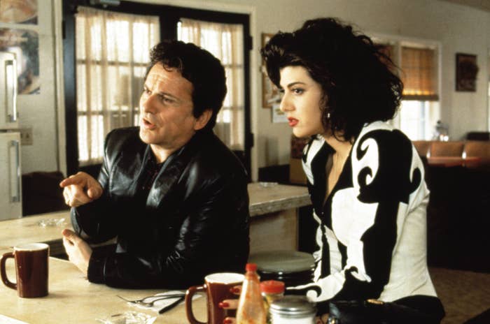 A man and a woman sit at a diner counter, engaged in conversation. The woman wears a patterned, fitted top; the man wears a leather jacket