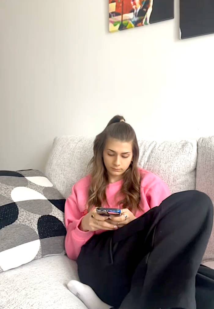 Person sitting on a sofa, looking at their phone, wearing a casual pink top and black pants, with abstract art pieces hung on the wall behind