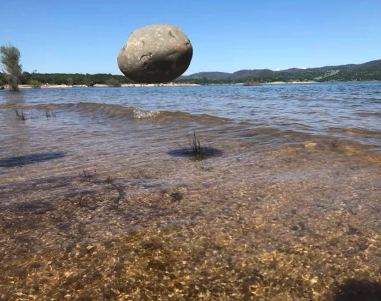 Large rock appears to hover above a serene lake with gentle waves and hills in the background