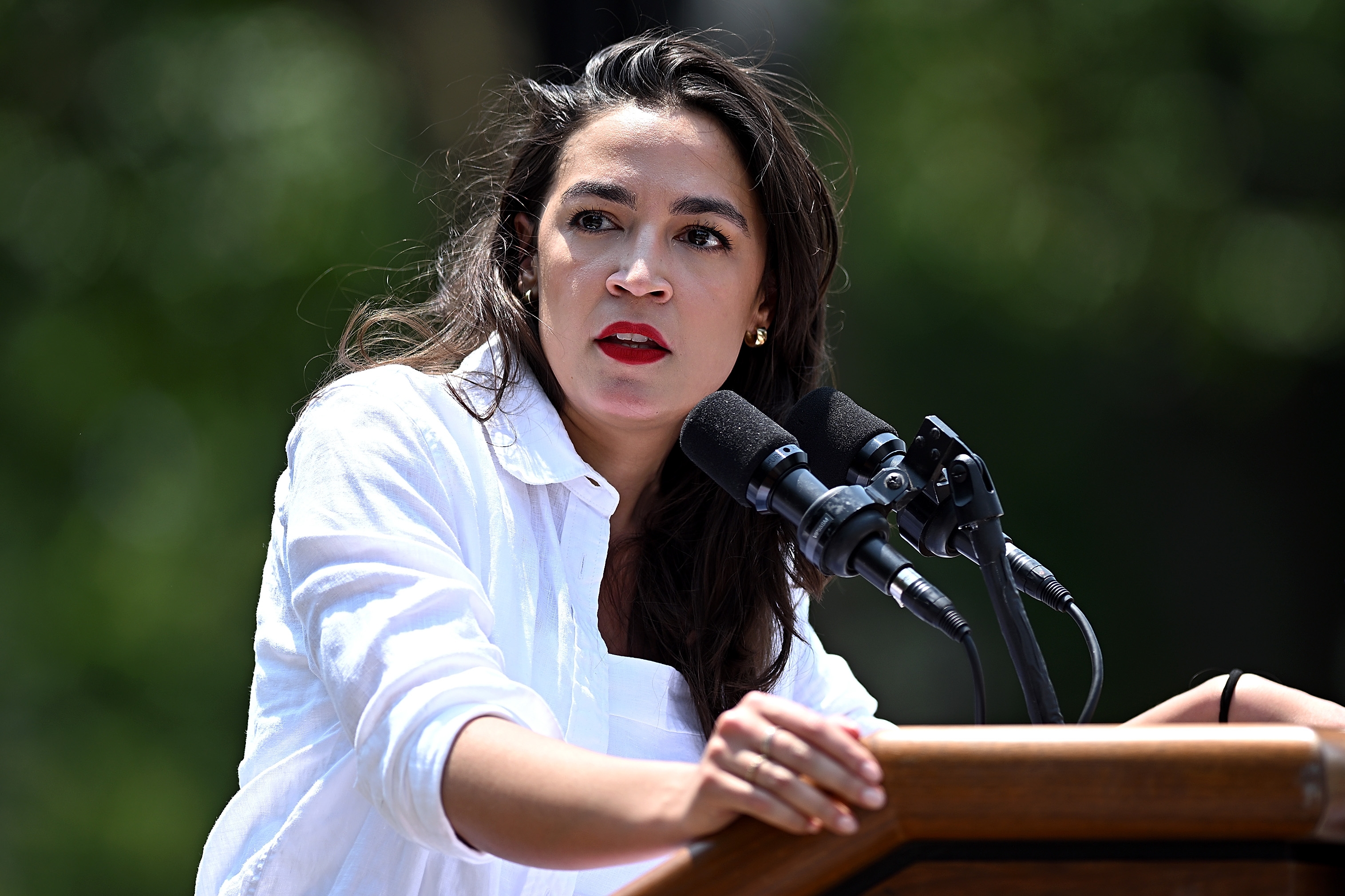 Person speaking passionately at an outdoor event, wearing a white shirt and standing at a podium with multiple microphones