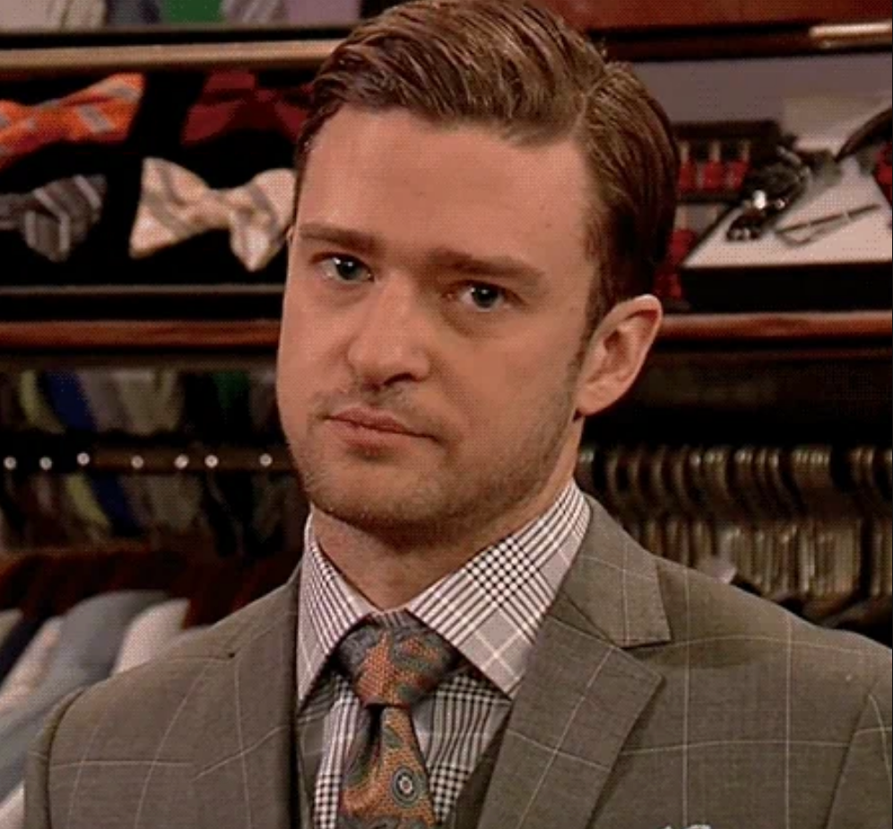 A person in a suit with a patterned dress shirt and tie stands in a closet with clothes on hangers behind them