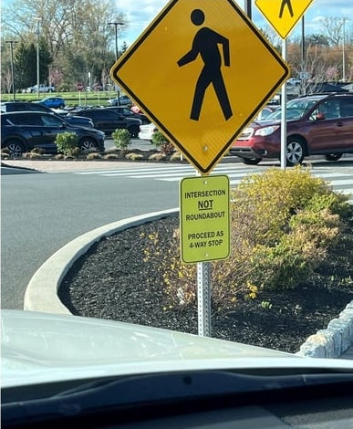 Pedestrian sign at a road. Small sign below reads: "Intersection NOT roundabout. Proceed as 4-way stop"