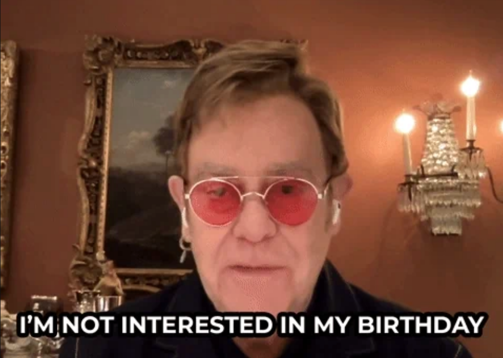 Elton John wearing round glasses, standing indoors, with text saying, "I'm not interested in my birthday"