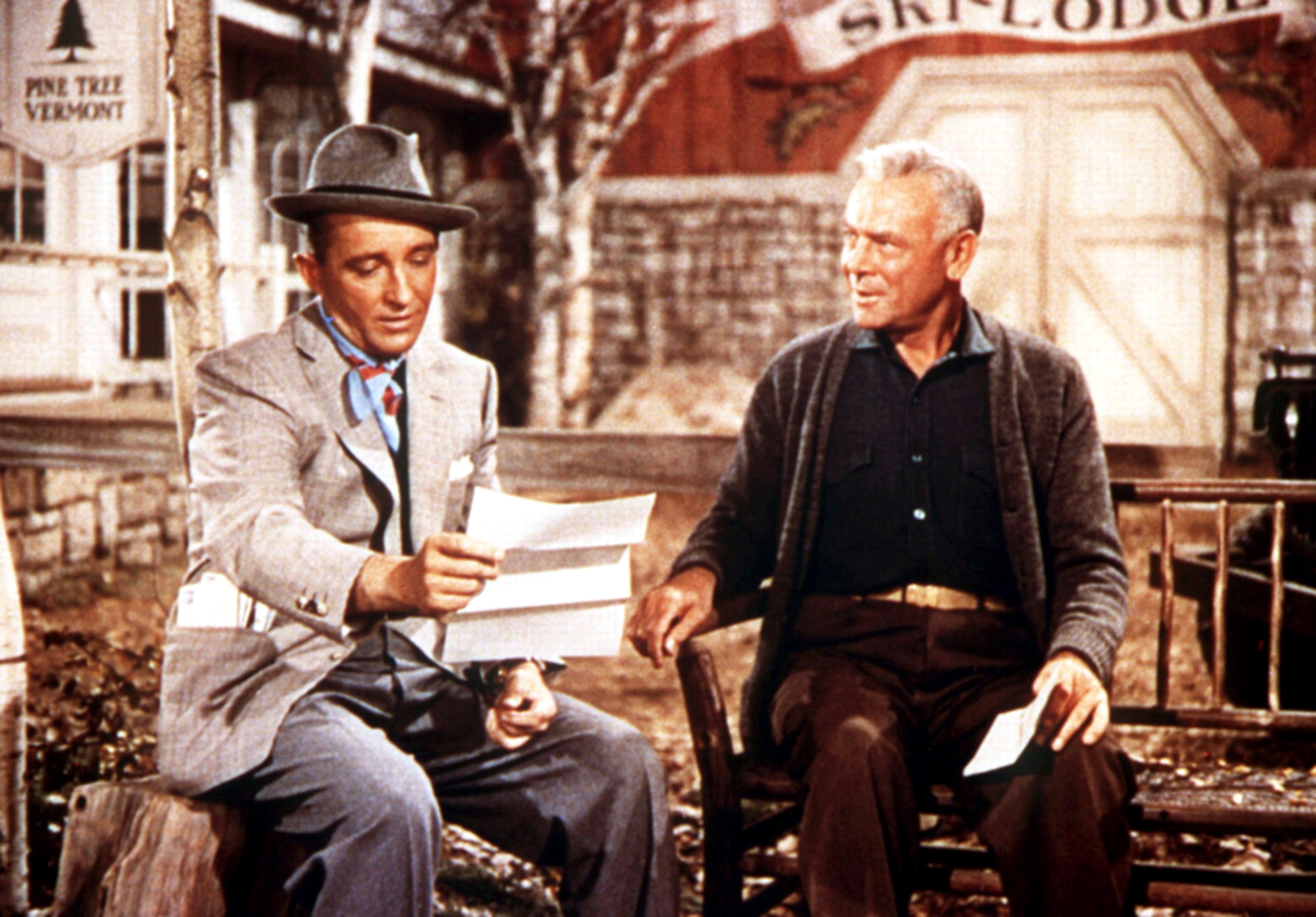 Two men in a rustic setting, one in a suit and hat, reading a letter. The other man wears a cardigan, sitting with a warm expression