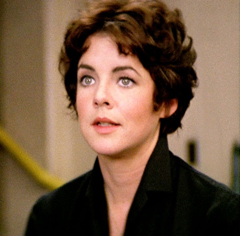 Person with short, dark hair wearing a collared jacket looks thoughtfully ahead indoors