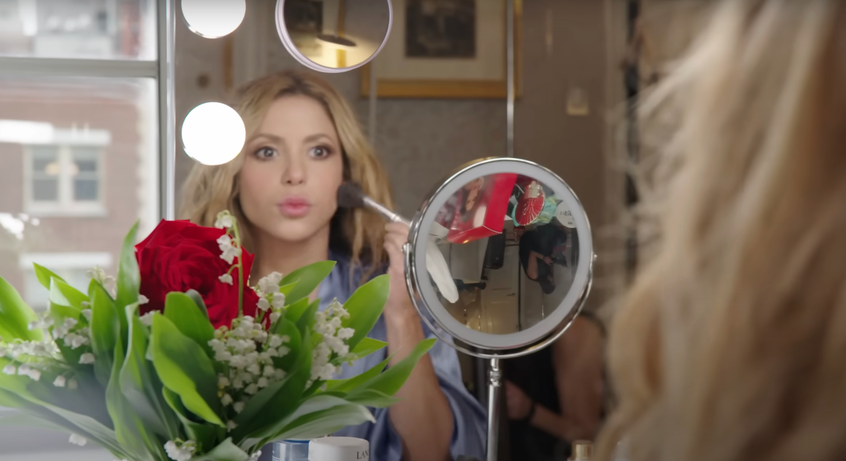 Shakira applying makeup at a vanity next to flowers, looking into a mirror with focus and precision