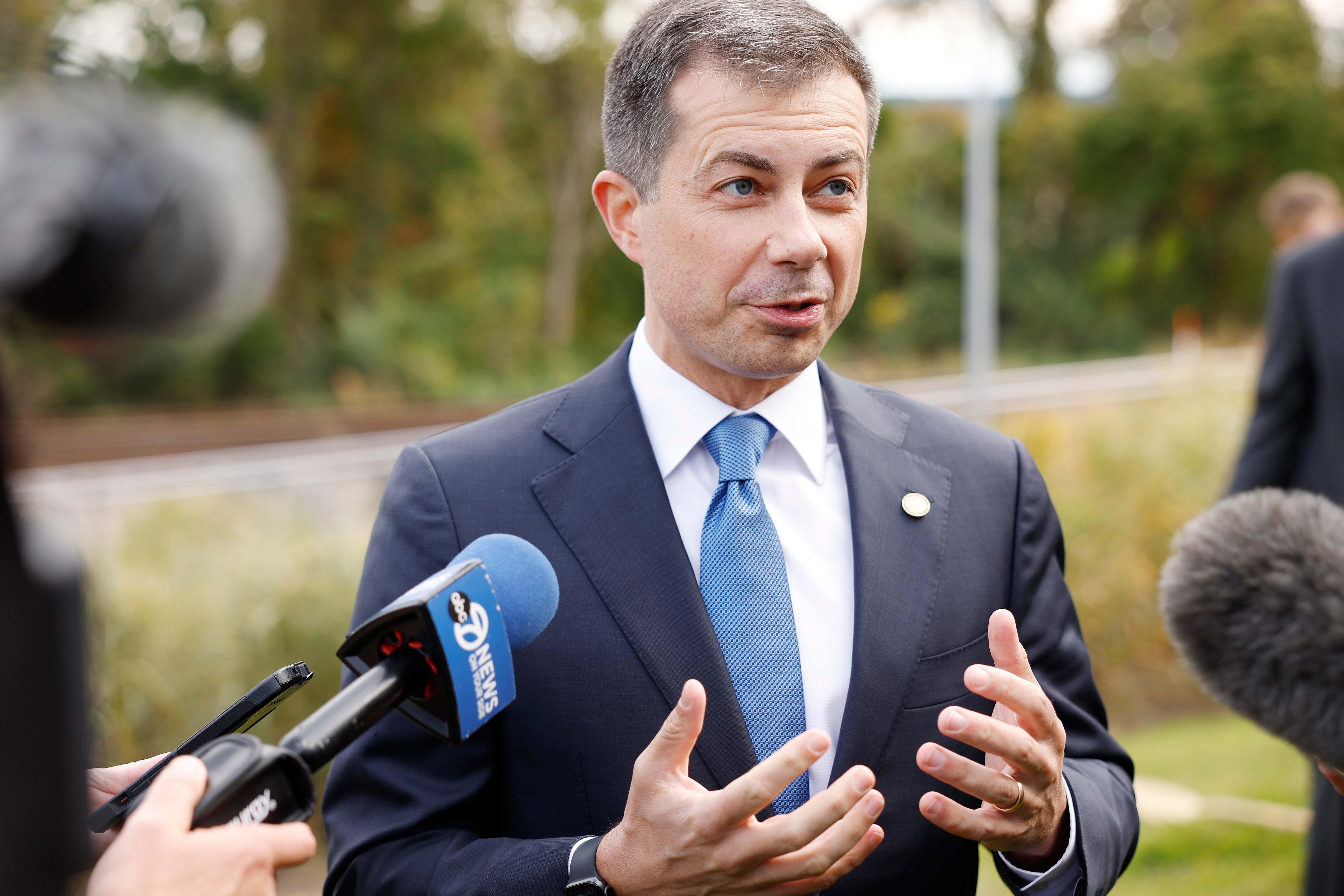 Person in a suit and tie speaks to reporters outdoors, gesturing with hands