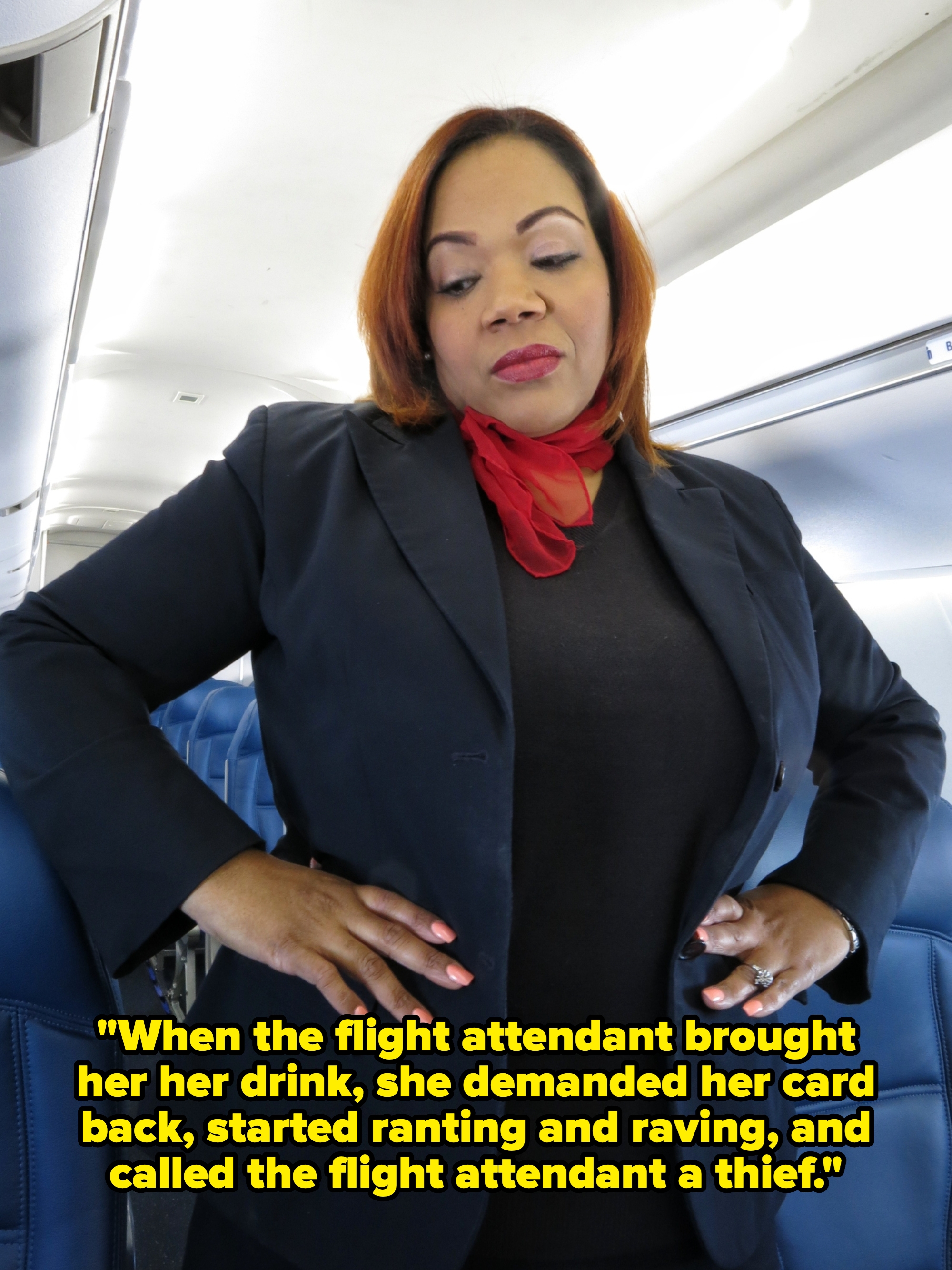 Person in an aircraft cabin wearing a black outfit with a red scarf, hands on hips, looking down contemplatively