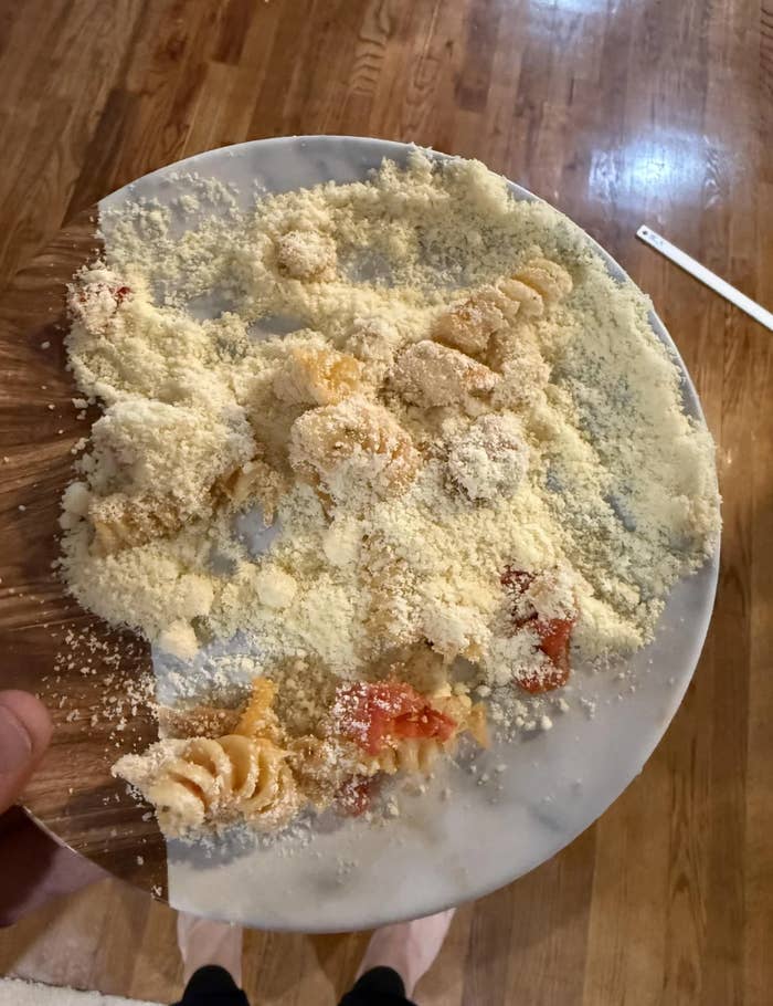 A plate of pasta accidentally flipped upside down, covered in grated cheese, with some pasta and sauce visible on the floor