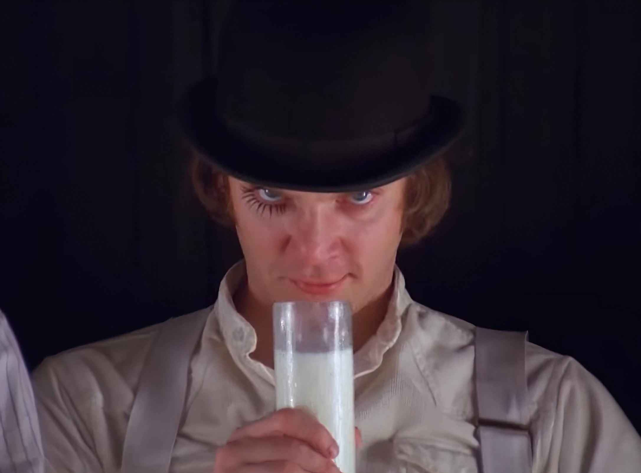 A person wearing a bowler hat and suspenders holds a glass of milk, looking intently at the camera