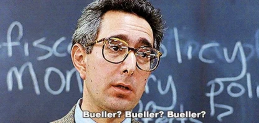 Ben Stein, a person with glasses in a tweed suit, stands in front of a chalkboard, saying "Bueller? Bueller? Bueller?" in a classroom setting