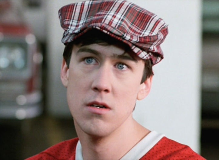 Person wearing a plaid cap and casual sweater looks up with a neutral expression, standing in front of a blurred vehicle