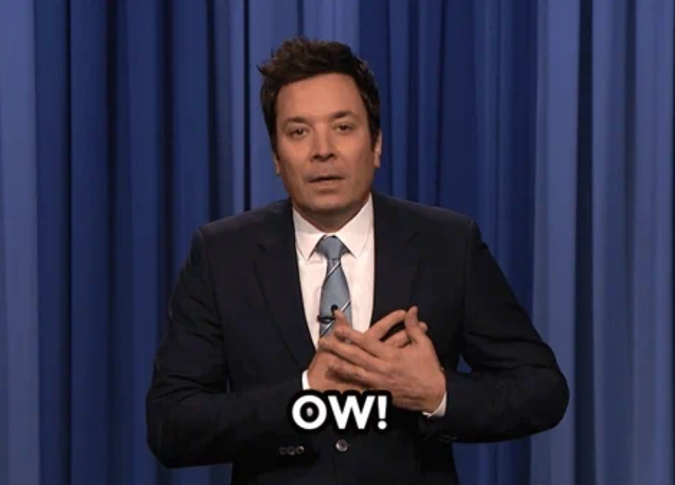 A person in a suit stands on stage, hands clasped over their chest with "Ow!" written below, suggesting surprise or mild discomfort