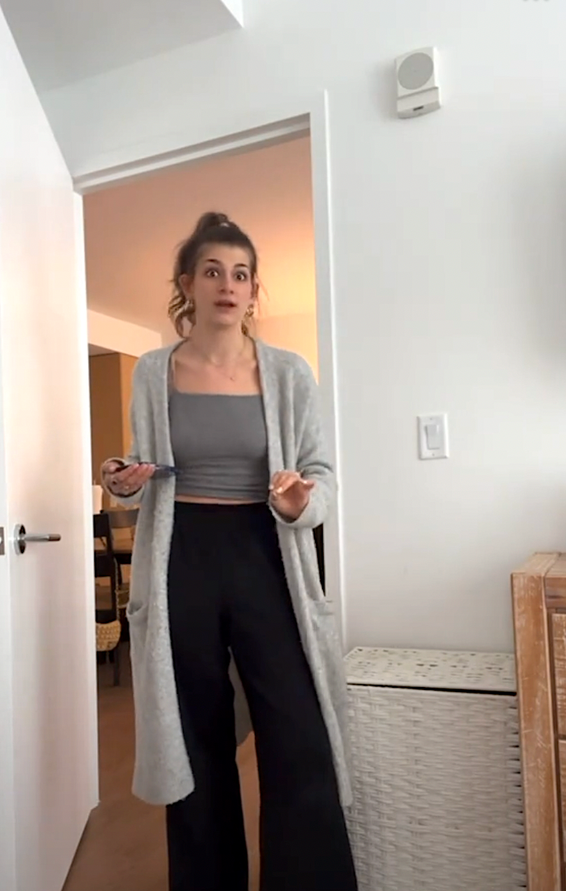 Person standing in doorway, wearing a long cardigan, crop top, and wide-leg pants, holding a phone and pen, with a surprised expression