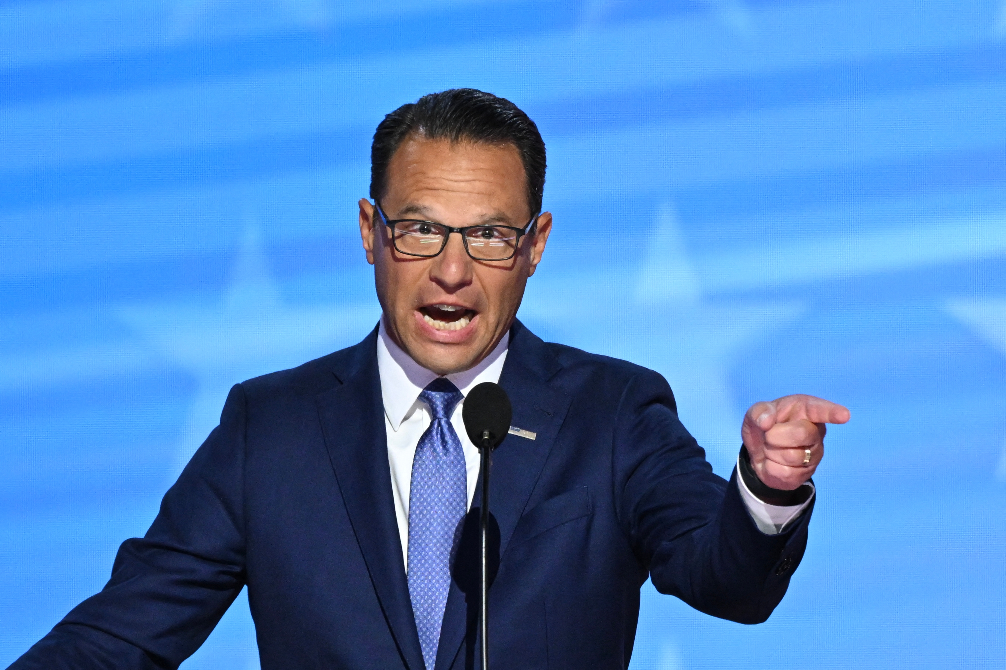 A man in a suit and tie speaks passionately at a podium, gesturing with his hand, against a backdrop with star motifs