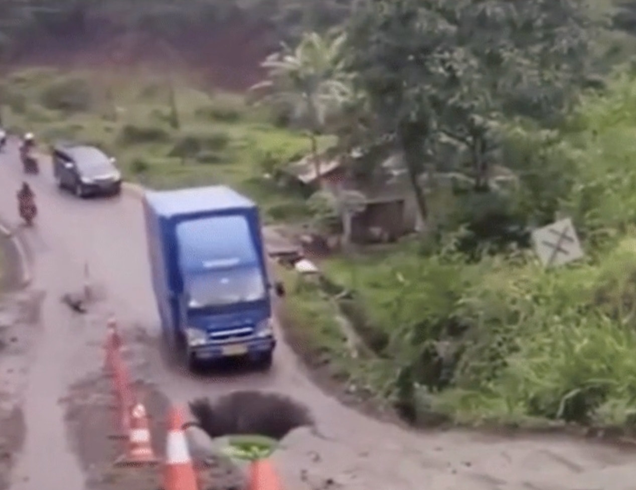 Large truck on a narrow, rural road maneuvering around a deep pothole beside a traffic cone. Passengers car in the background