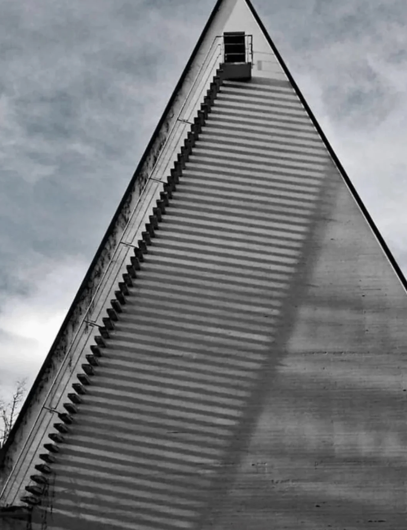 Triangular building facade with a staircase casting dramatic shadows. A small platform is visible at the top