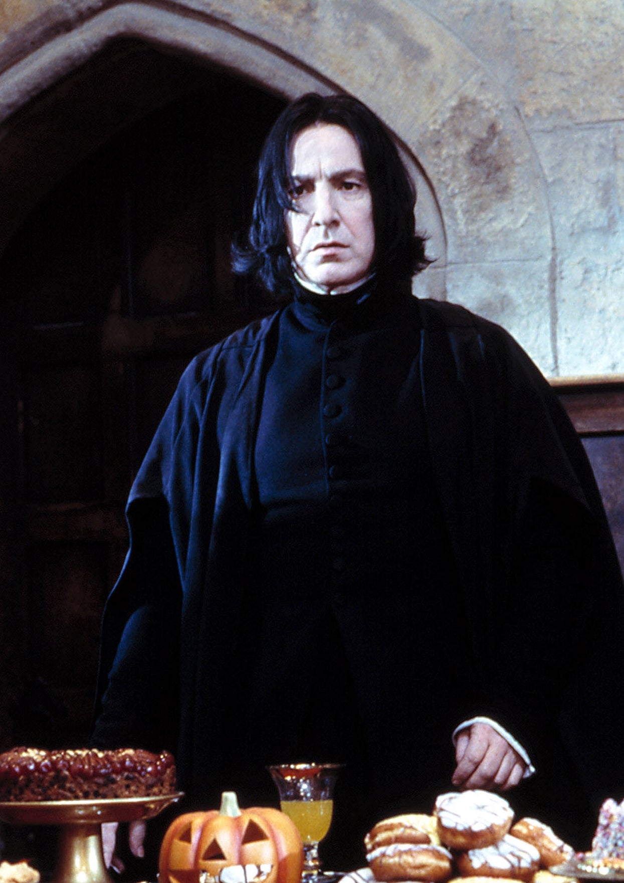 Person dressed in dark robes stands behind a table with various desserts, including a cake and a pumpkin container, in a stone room