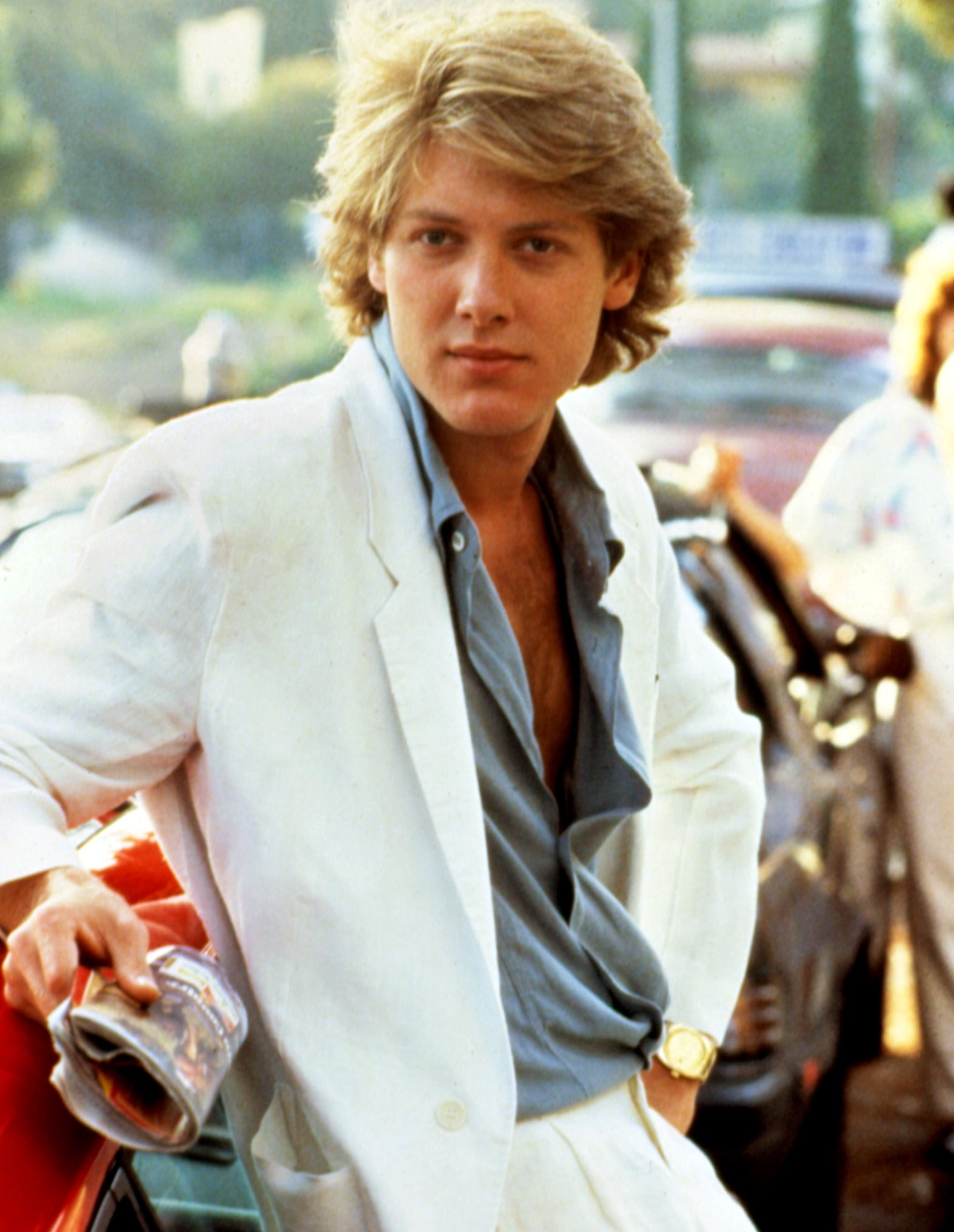 Person in a casual suit leans on a car, looking at the camera. Their style features an open-collared shirt and a relaxed pose