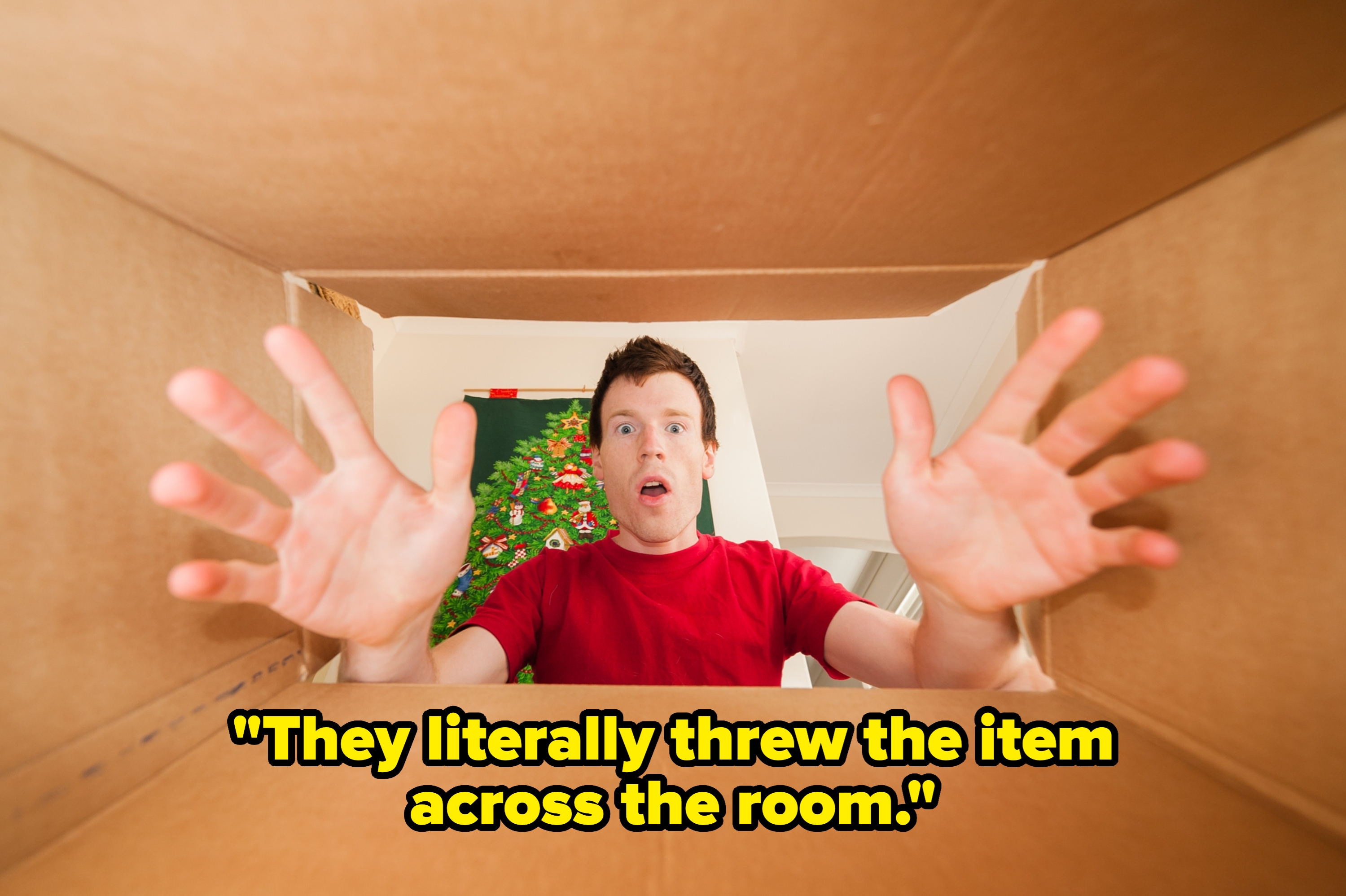 Man in red shirt inside large box, looking surprised with hands raised. Seen from the box's perspective. Background has indoor decorations