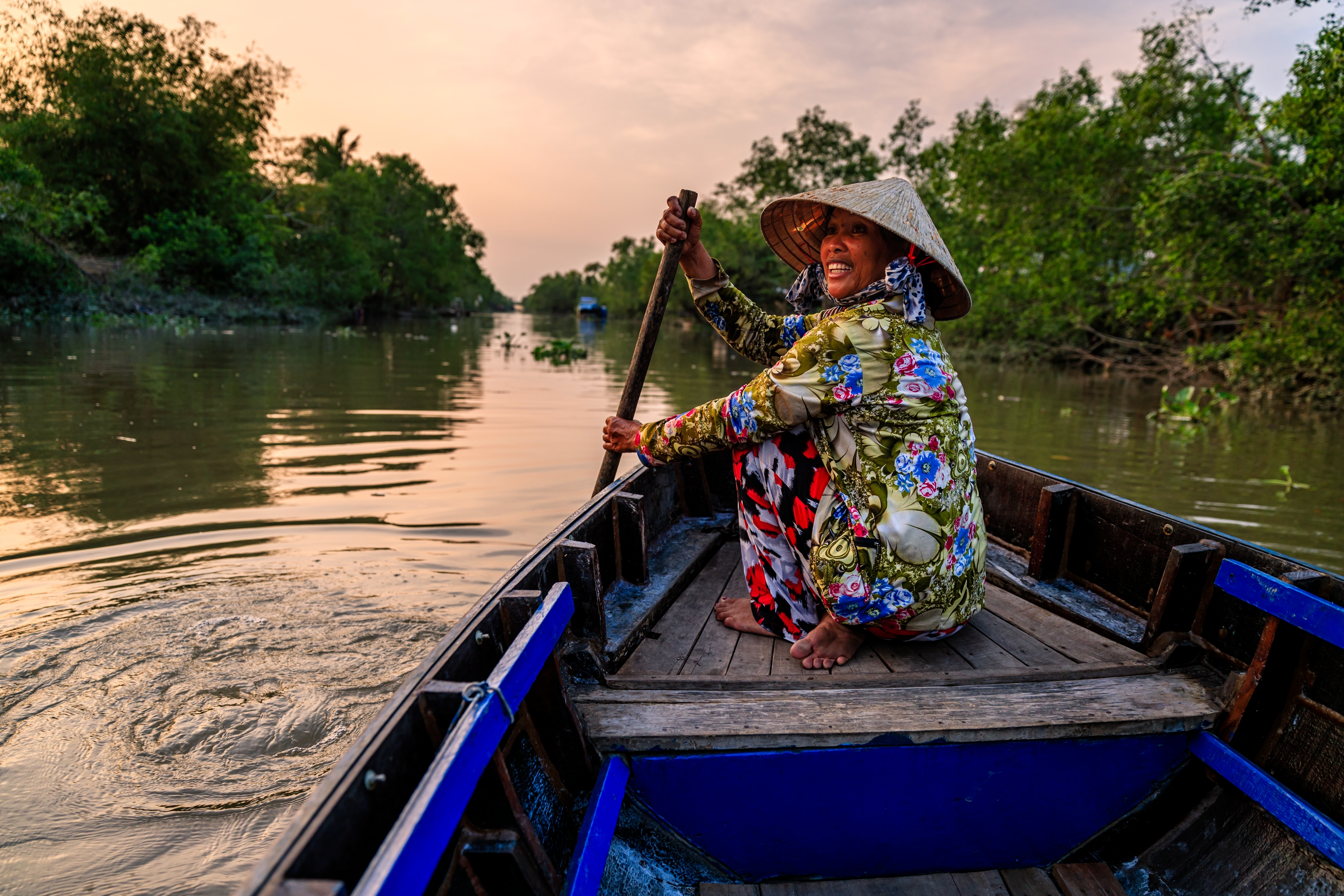 Person paddling a small boat on a serene river, wearing a traditional conical hat and floral clothing, surrounded by lush greenery