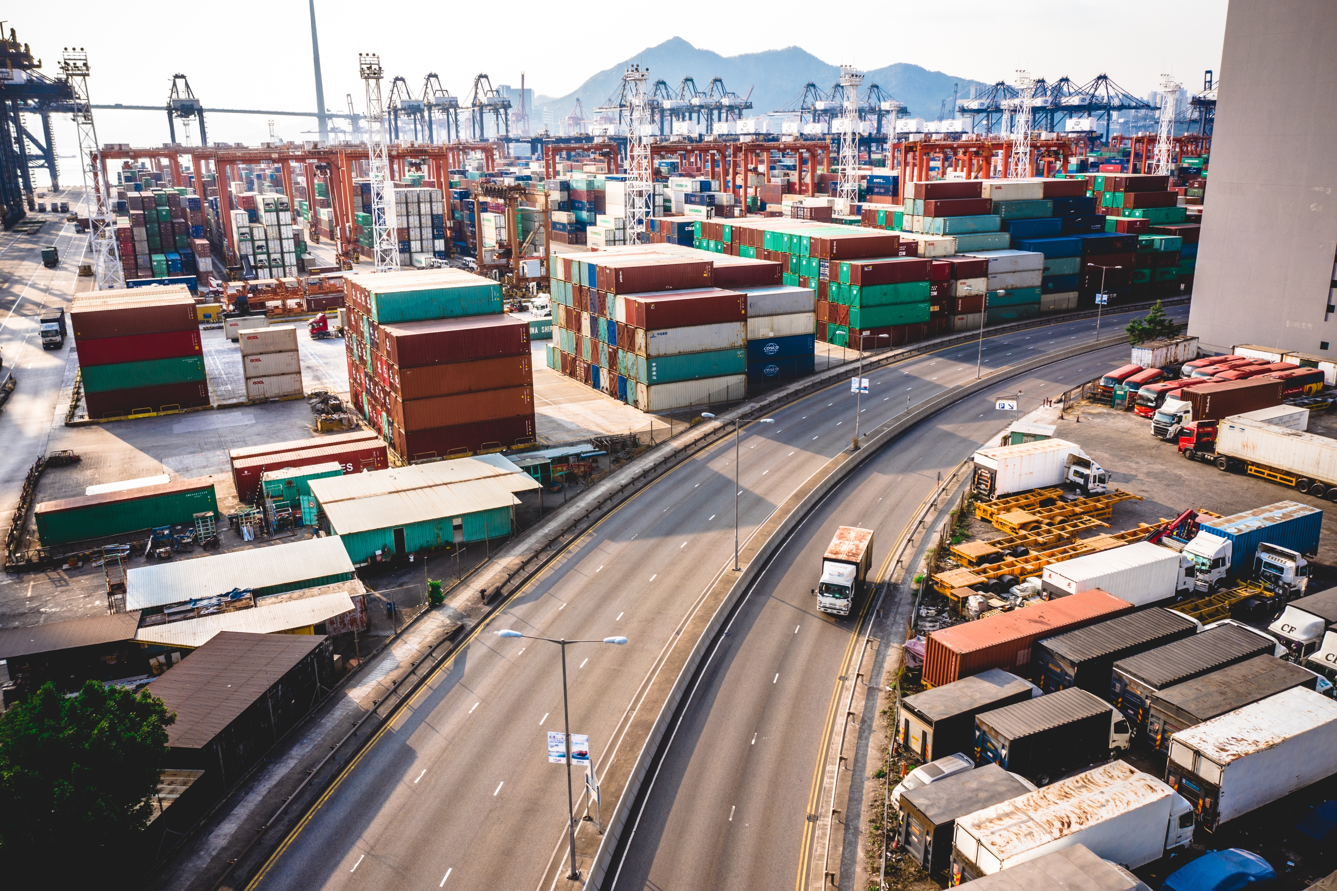 Aerial view of a bustling port with stacked shipping containers, cranes, and trucks, set against a backdrop of mountains, illustrating global trade