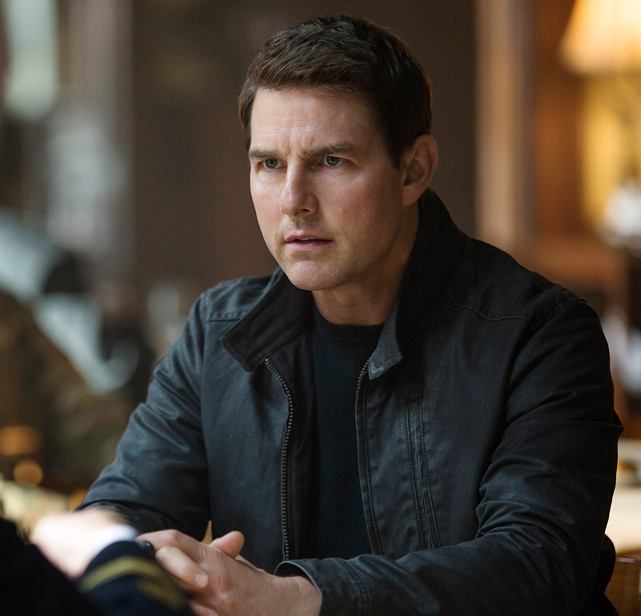 A person in a casual jacket sits at a table, engaged in conversation with a military personnel in uniform, appearing focused and attentive