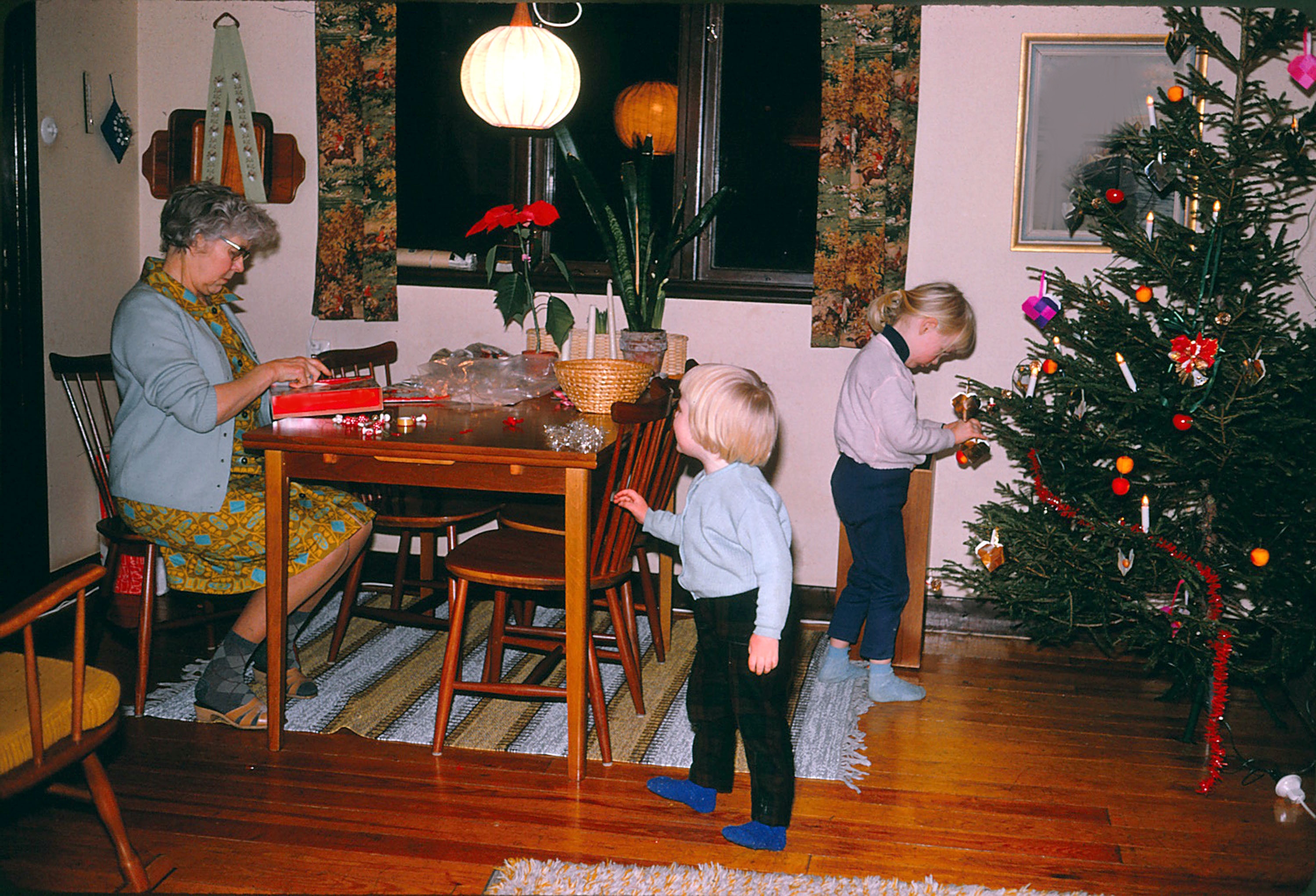 A woman wraps gifts at a dining table while two children decorate a Christmas tree in a cozy living room