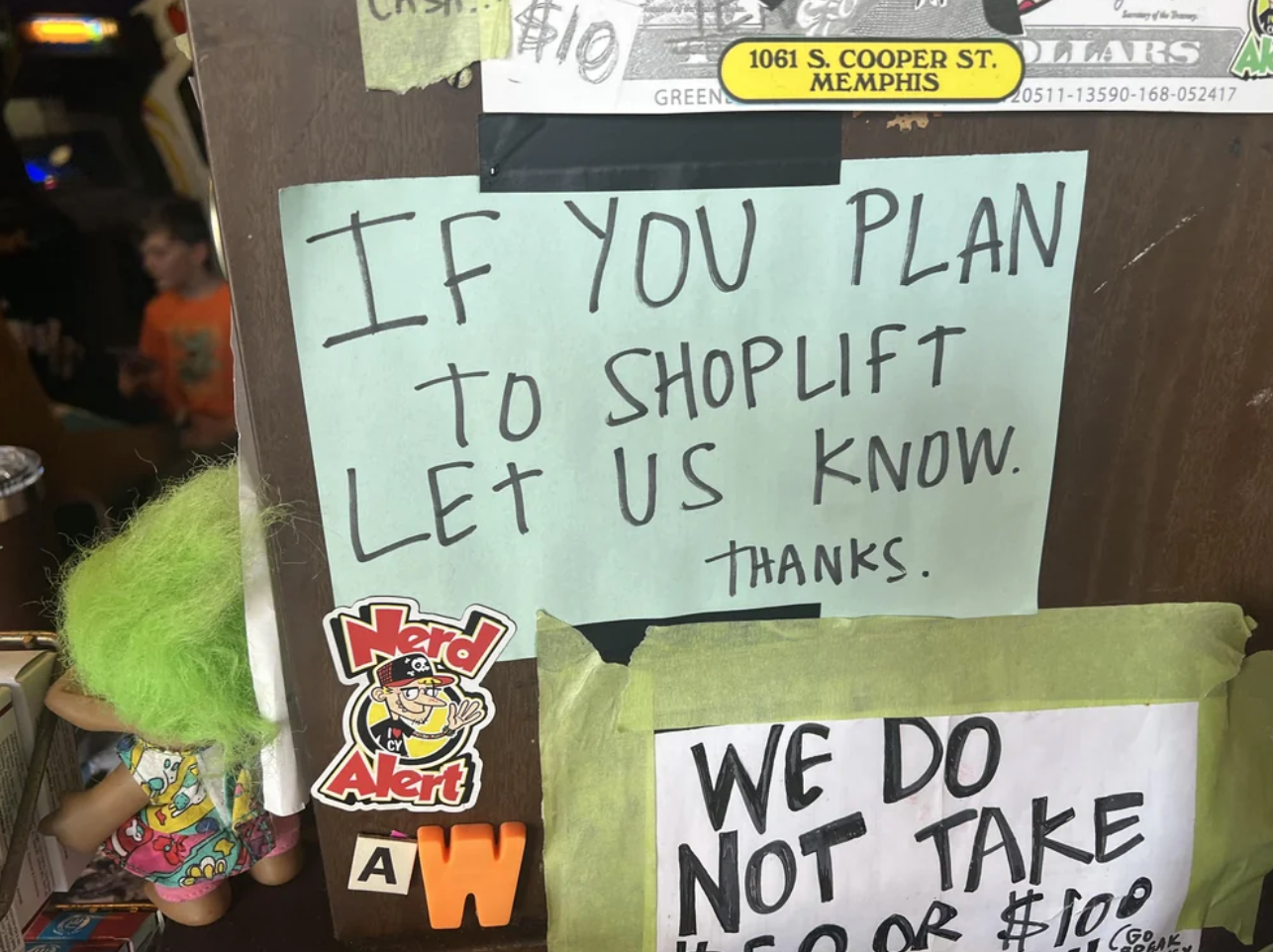 Sign reads: "If you plan to shoplift, let us know. Thanks." Nearby, a troll doll and stickers are visible