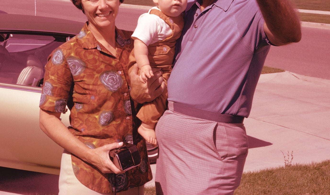 A man pointing enthusiastically, holding a baby, stands next to a smiling woman by a car in a suburban neighborhood