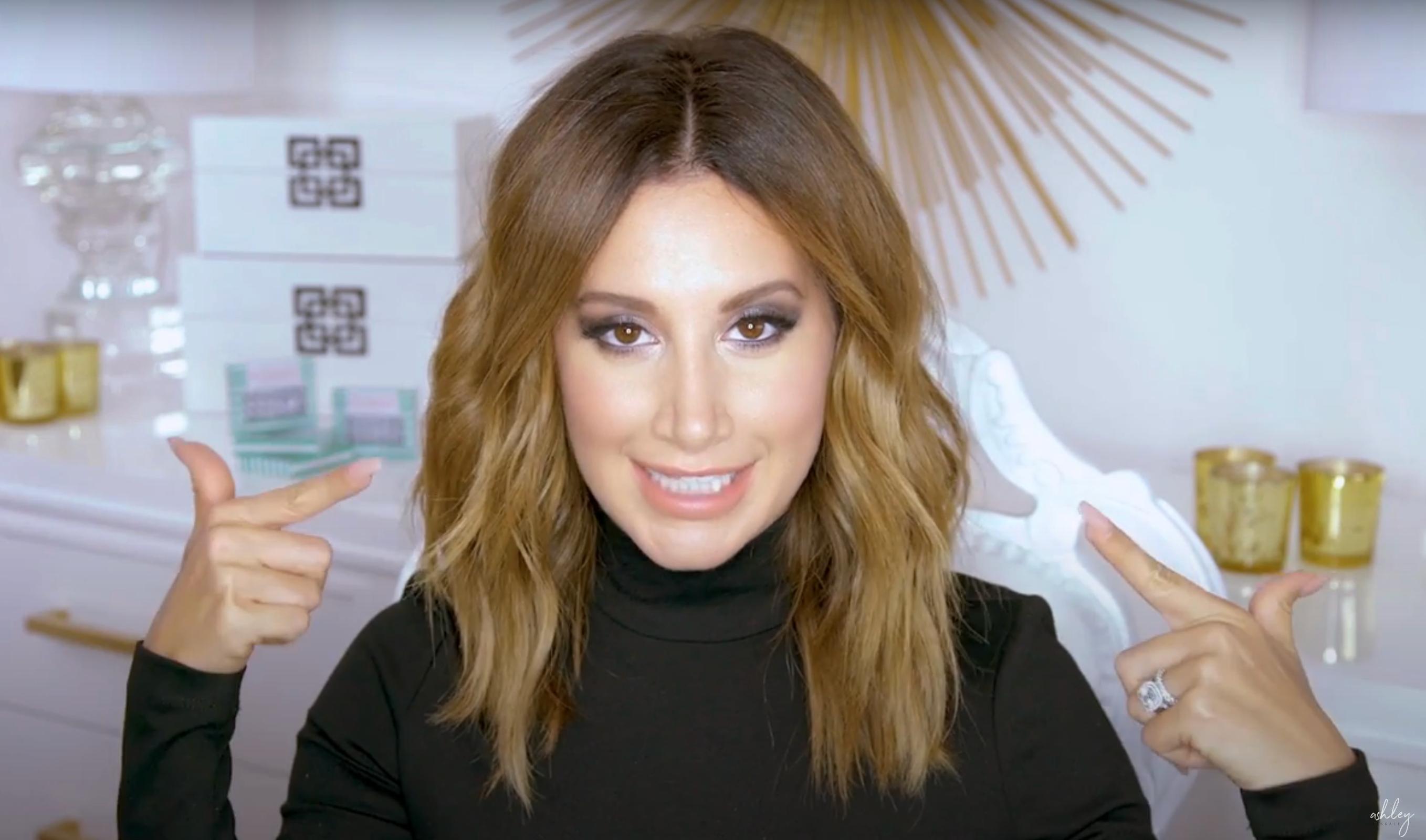Ashley pointing at her makeup look, which is bold with dark eyeshadow and heavy liner but light lipstick
