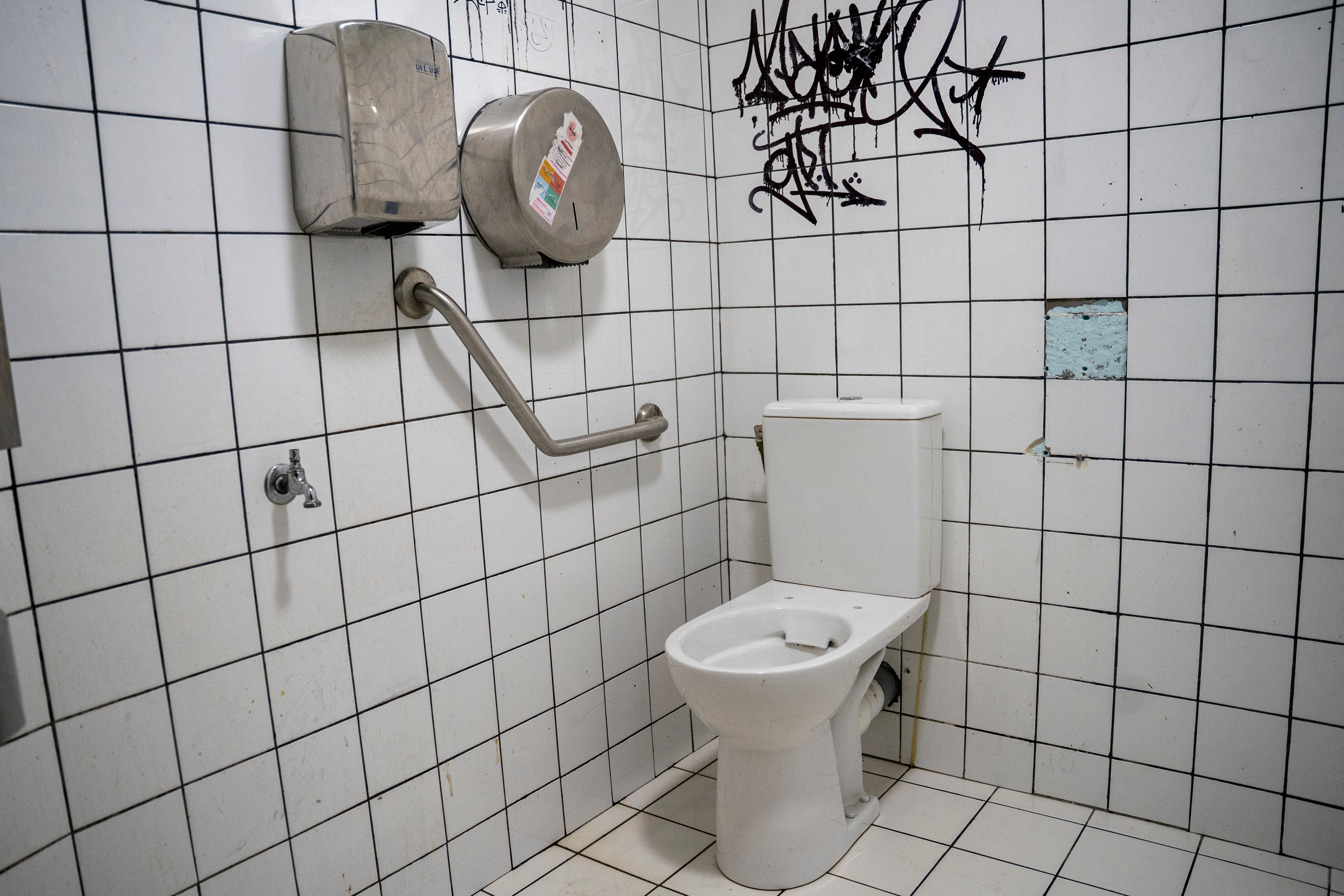 Accessible public restroom with a handlebar next to the toilet, graffiti on tiled walls