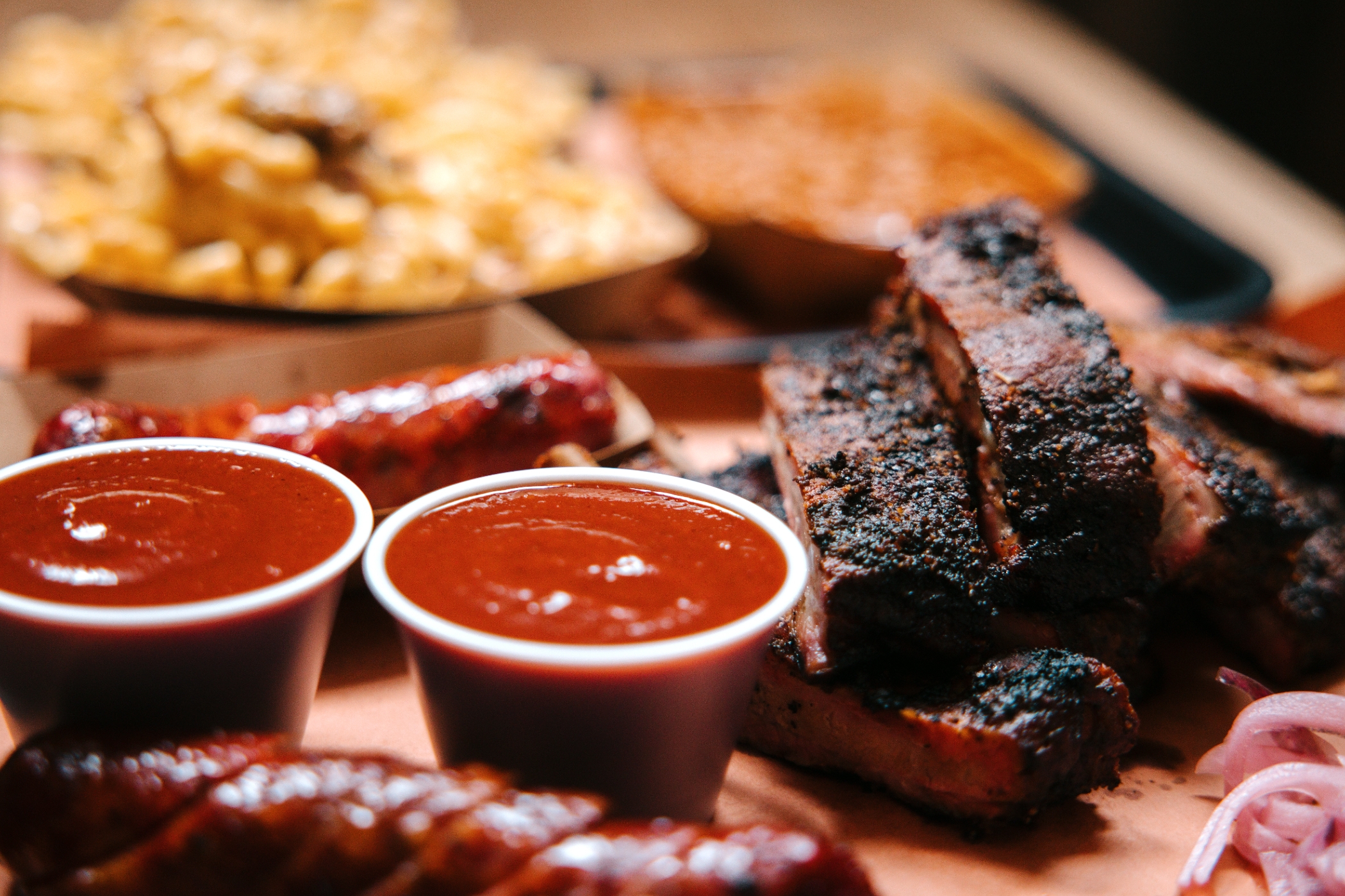 Barbecue spread featuring ribs, sausages, and two cups of sauce, with a side of macaroni and cheese in the background. Ideal for food travel articles