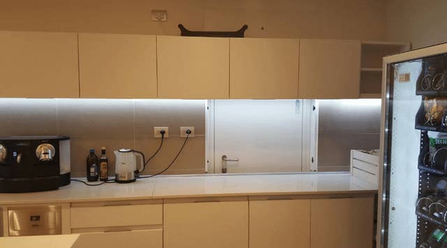 Modern kitchen with white cabinets, a coffee maker, electric kettle, and various bottles on the counter. A refrigerator with glass door is on the right