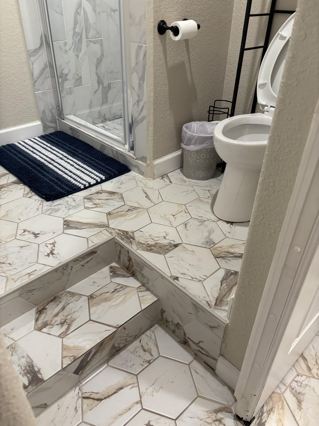 Bathroom with hexagonal marble-patterned tiles. Two steps lead to an elevated area with shower and toilet, which is tucked right next to the steps