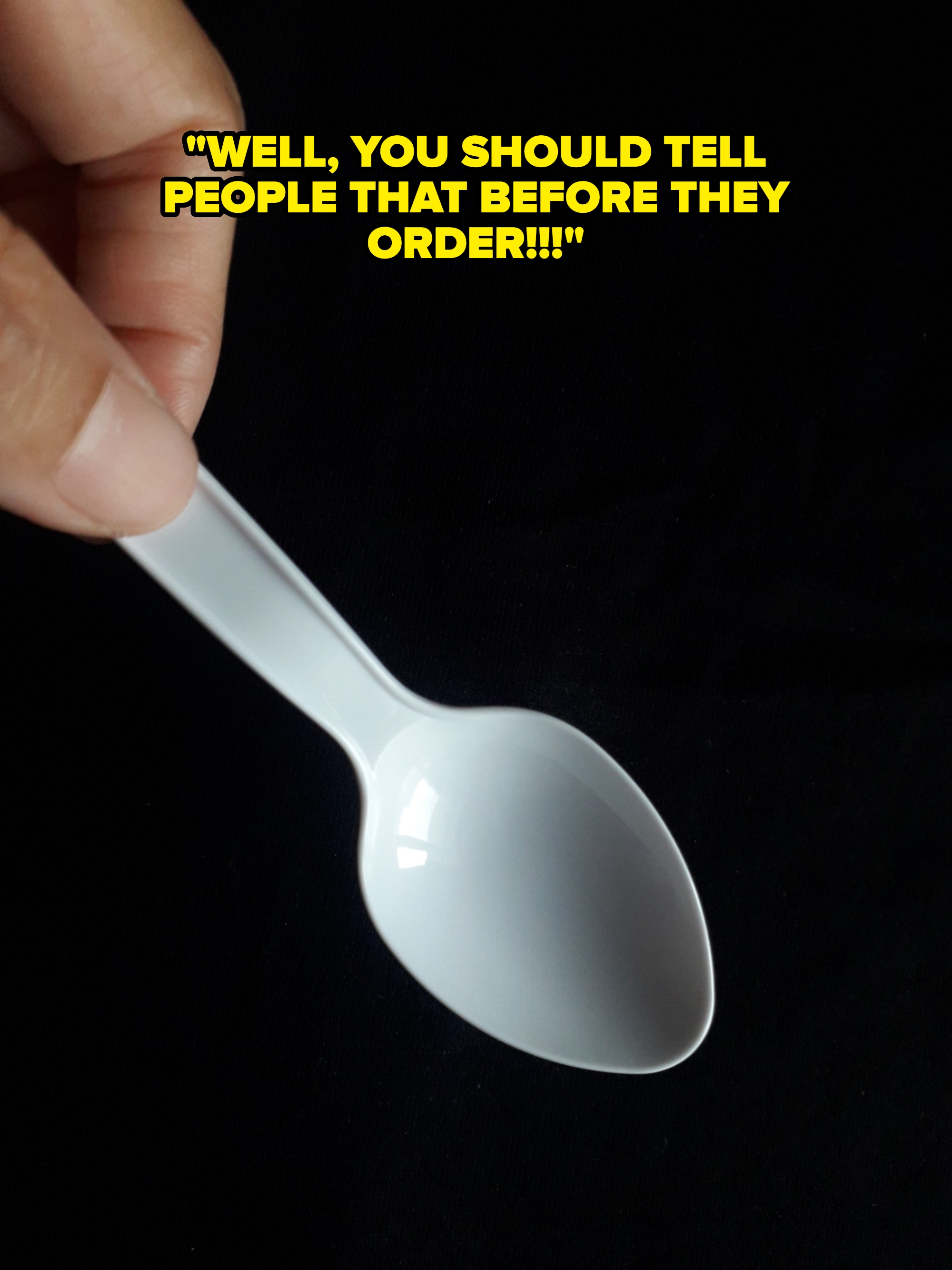 A hand holding a small white plastic spoon against a dark background