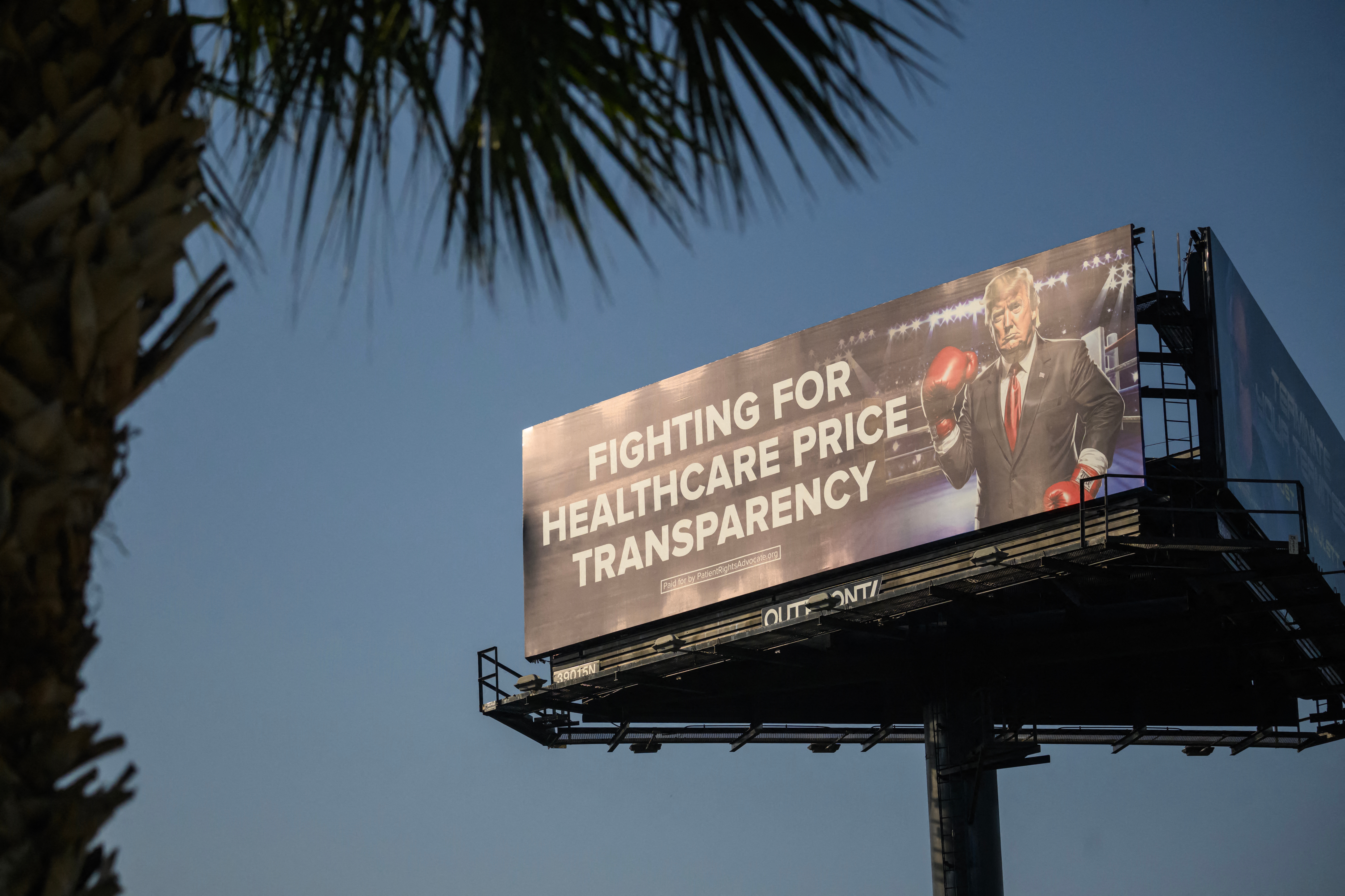 A billboard shows a man in a suit with boxing gloves, promoting healthcare price transparency. A palm tree is partially visible
