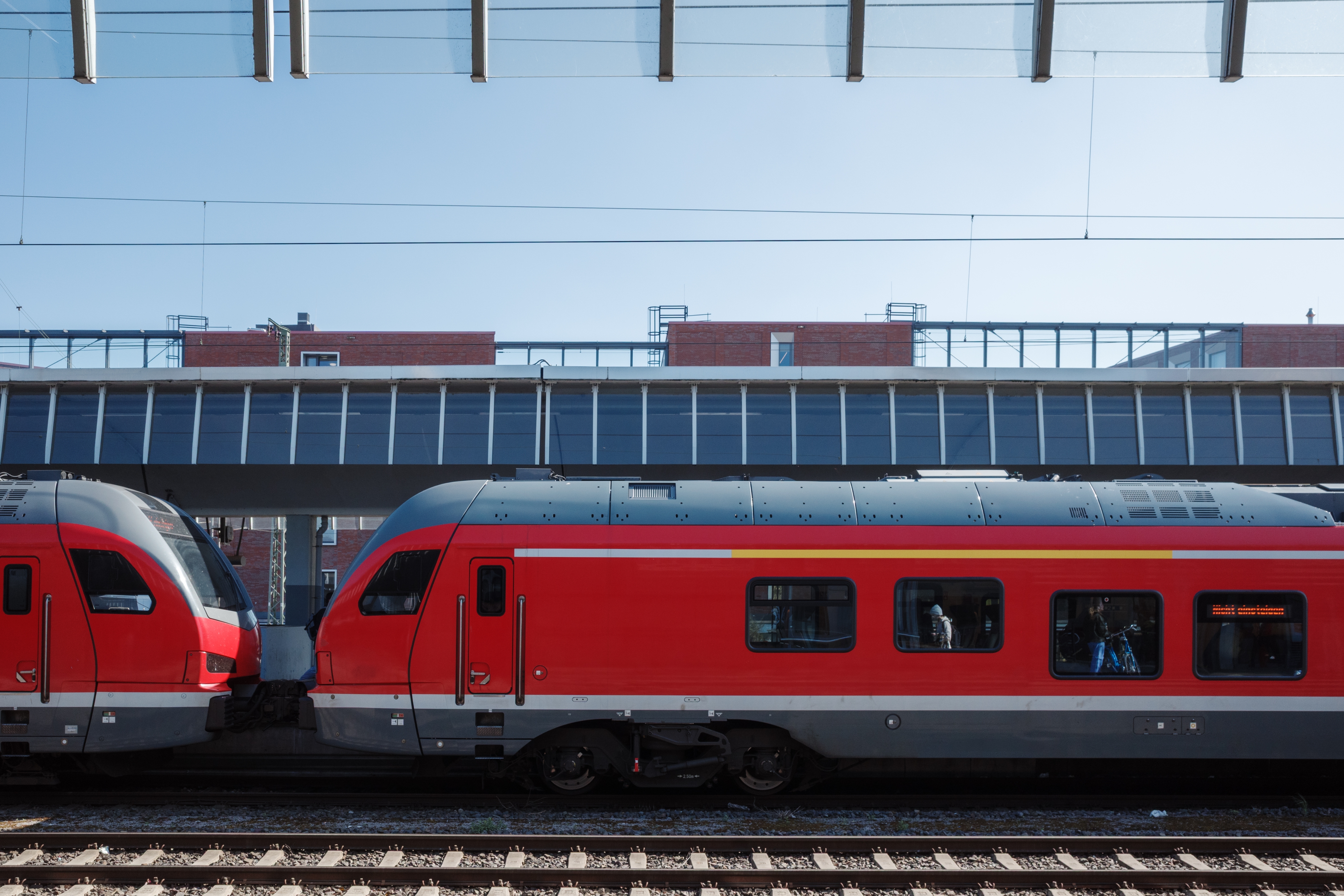 Two modern trains are stopped at a station, with passengers visible through the windows. The setting is urban, beneath a covered platform